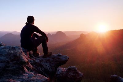 Adult tourist in black trousers, jacket and dark cap sit on cliff's edge and looking to misty hills