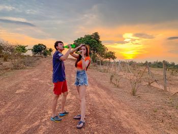 Couple drinking beer while standing on dirt road during sunset