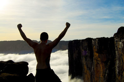 Rear view of shirtless man celebrating success while standing on mountain against sky