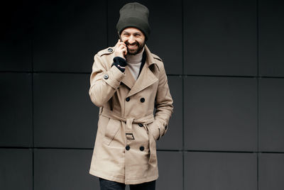 Portrait of smiling man talking on phone while standing against wall