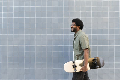 Happy man walking with skateboard in front of tiled wall