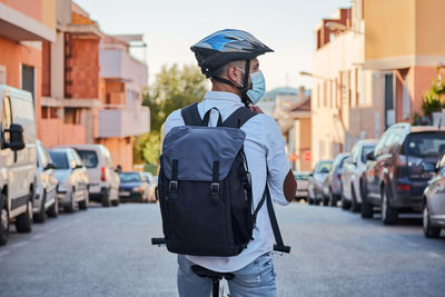 Back view of unrecognizable male in helmet and protective mask with backpack riding bike on city street while commuting to work