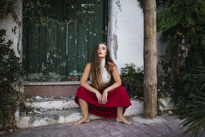 Side view of charming woman in skirt and crop top sitting on stone steps in patio with green plants and looking at camera