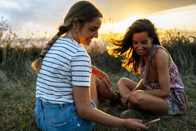 Young women smiling woman sitting against sky during sunset