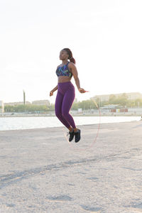 Full body fit black woman athlete in sportswear jumping with rope at the sea shore