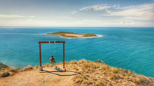 Rear view of man sitting on swing by sea against sky