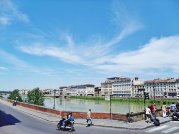 People on street by canal and buildings against cloudy sky during sunny day