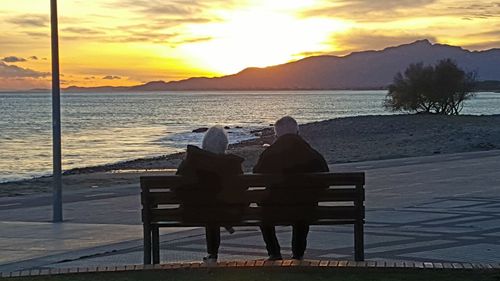 Rear view of people sitting on beach against sky during sunset