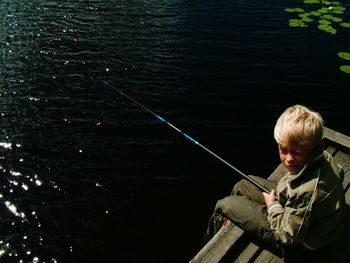 High angle view of boy fishing by lake while sitting on pier