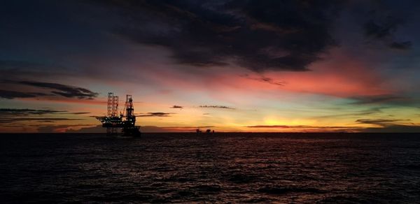 Silhouette oil factory on sea against dramatic sky during sunset