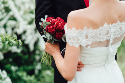 Midsection of bride and groom dancing affectionately with bunch of red flowers