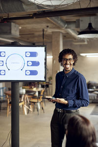 Happy businessman holding digital tablet while standing by television set during meeting in office