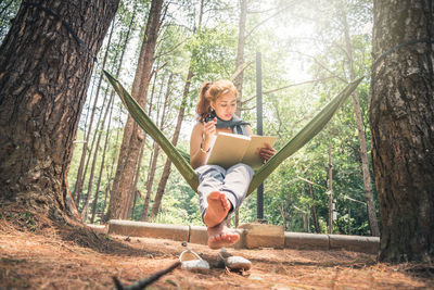 Full length of woman reading book while sitting in hammock at forest