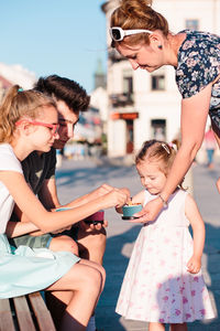 Mother giving food to children in city during sunny day