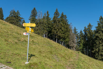 Road sign by trees on field against clear blue sky