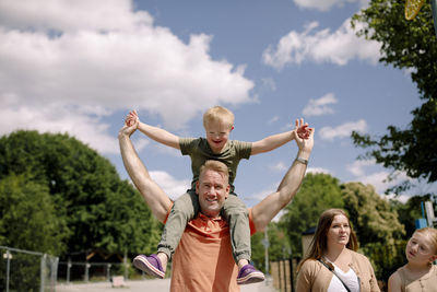 Father carrying son on shoulders while walking by woman and daughter at park during sunny day