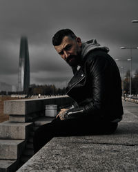 Side view of young man sitting on retaining wall against sky