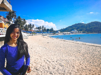 Portrait of young woman standing at beach against blue sky