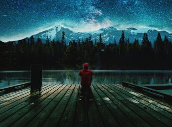 Rear view of man sitting on pier over lake against sky at night