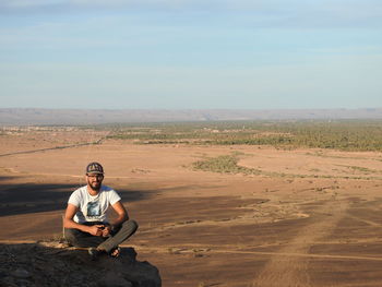 Portrait of young man sitting on desert against sky