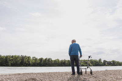 Back view of senior man standing with wheeled walker at riverside