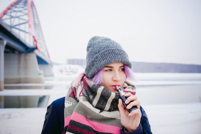 Young woman smoking while standing by bridge against sky during winter