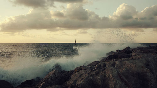 Waves splashing on rock against cloudy sky