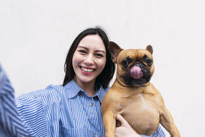 Happy woman carrying dog sticking out tongue in front of wall