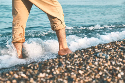 Male legs walking on pebble beach along the shore near the water with waves, low section.