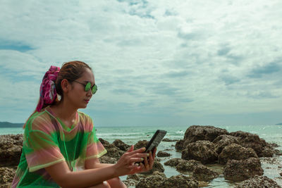 Woman using digital tablet while sitting at beach against sky