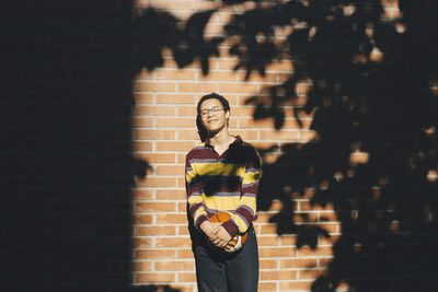 Smiling man with eyes closed holding soccer ball against brick wall