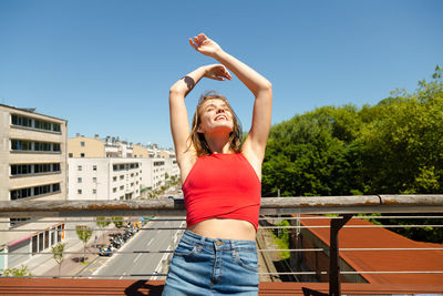 Smiling woman with arms raised standing against clear sky