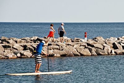 Rear view of men standing on rock by sea against clear sky