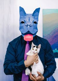 Man with origami paper mask in form of french bulldog holding kitten in room