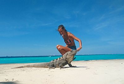 Side view portrait of young woman with reptile crouching at beach against blue sky during sunny day