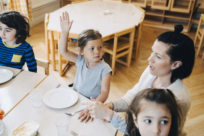 High angle view of girl with arm raised sitting amidst teacher and friends at table in classroom