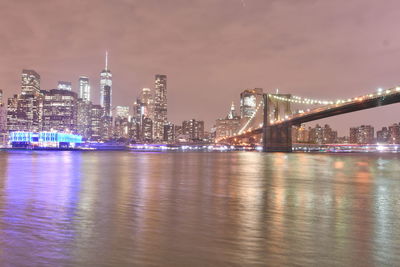 Illuminated modern buildings by river against sky at night