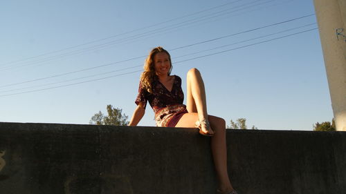 Low angle view of smiling young woman sitting on retaining wall against clear blue sky during sunny day