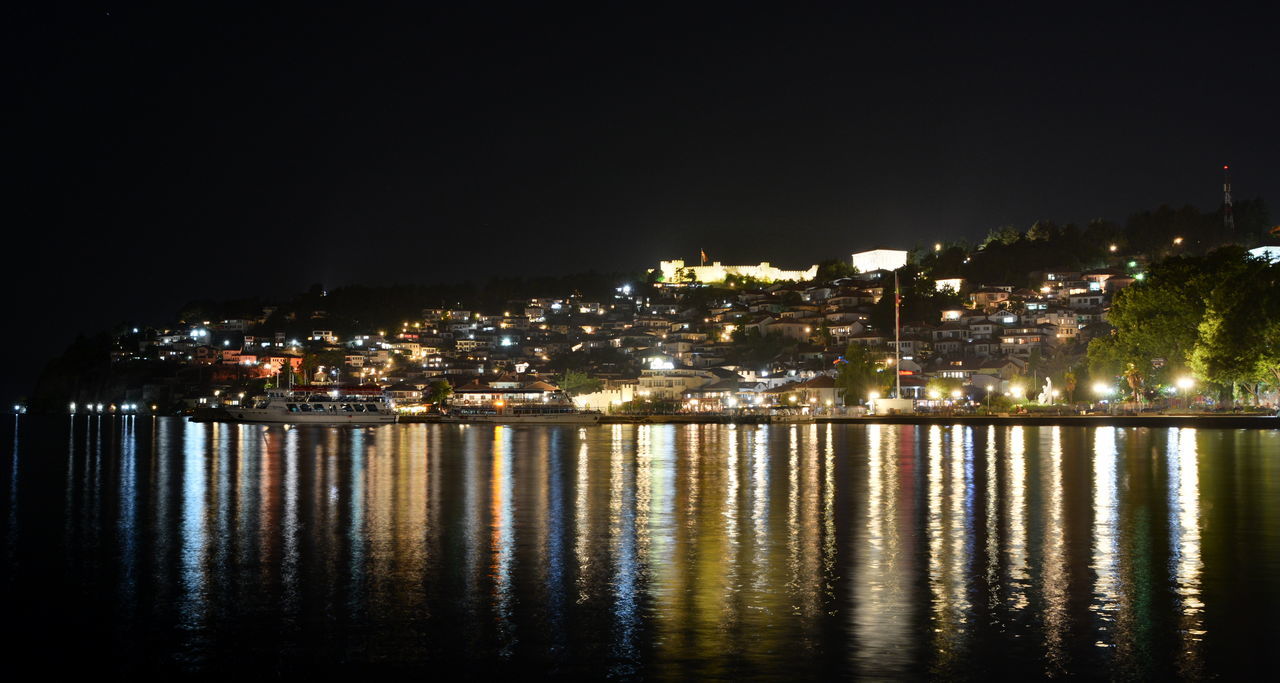 ILLUMINATED CITY BY SEA AGAINST SKY AT NIGHT