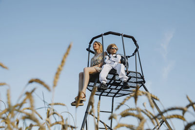 Son wearing space suit sitting with mother on lookout tower at field