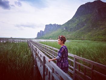 Thoughtful woman standing on wooden footbridge over grassy field against sky