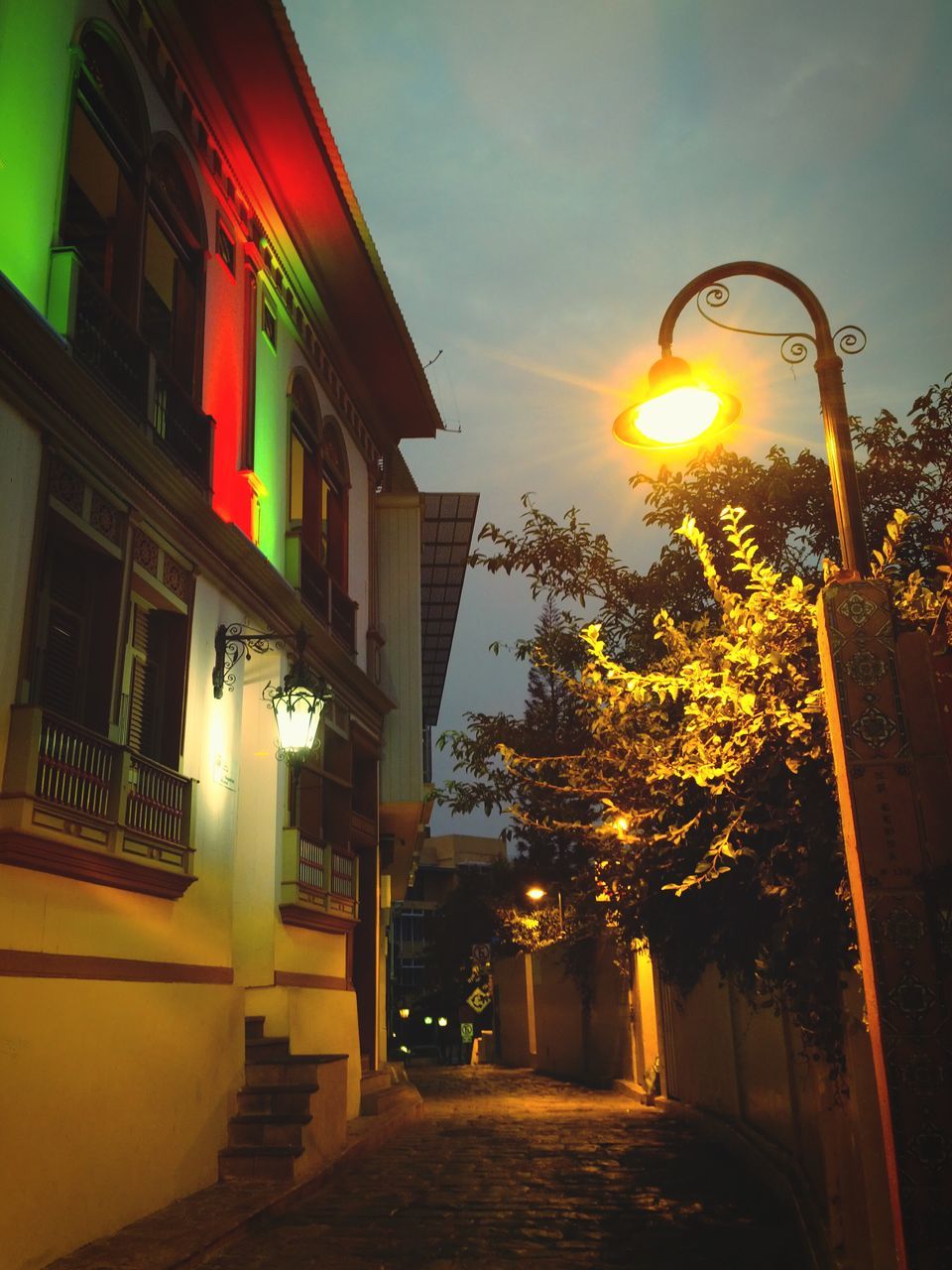 architecture, built structure, building exterior, street light, lighting equipment, sky, illuminated, sunset, the way forward, street, low angle view, tree, orange color, cloud - sky, sunlight, residential building, lantern, sun, dusk, no people