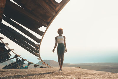 Full body of slim thoughtful african american female in trendy outfit and high heels walking near shabby wooden construction ind desert area on summer day