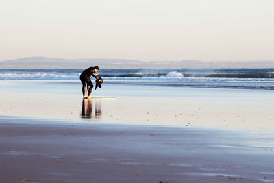 Man holding helmet while bending on sea shore against clear sky