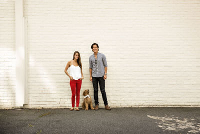 Portrait of couple with poodle standing against white brick wall