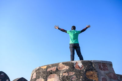 Low angle view of man standing on rock against clear blue sky