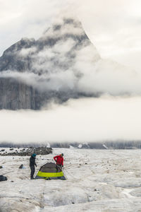 Two men set up a tent below a dramatic mountain peak.