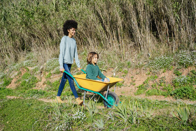 Father walking on a dirt track, pushing wheelbarrow, with his son sitting in it