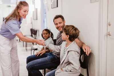 Smiling female pediatrician handshaking with boy sitting with family in hospital corridor