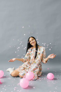 A pretty young woman sits near pink balloons and throws up confetti and smiles 